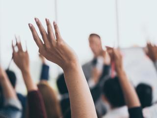 Hands raised in a classroom setting, with a blurred speaker in the background.