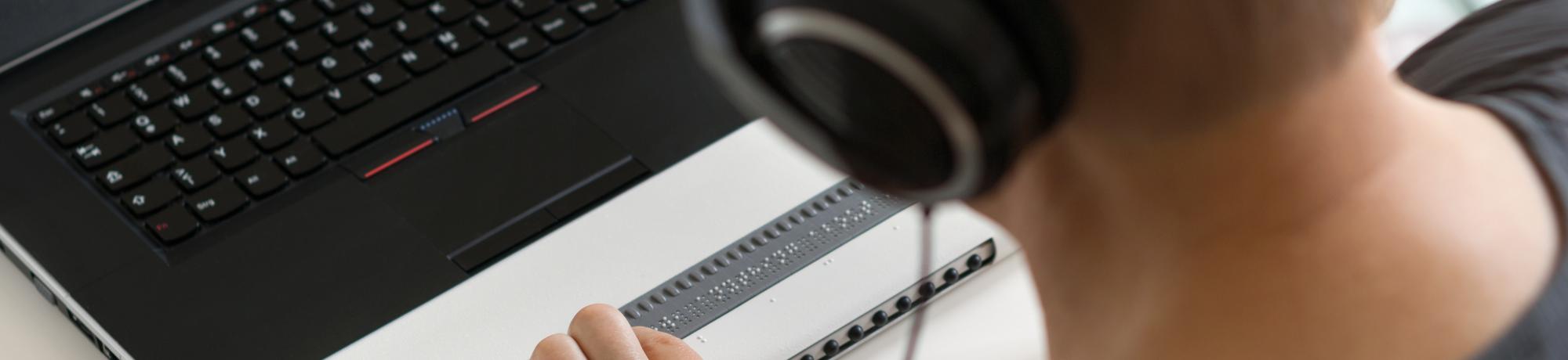 A person wearing headphones uses a laptop with an accessible Braille display.